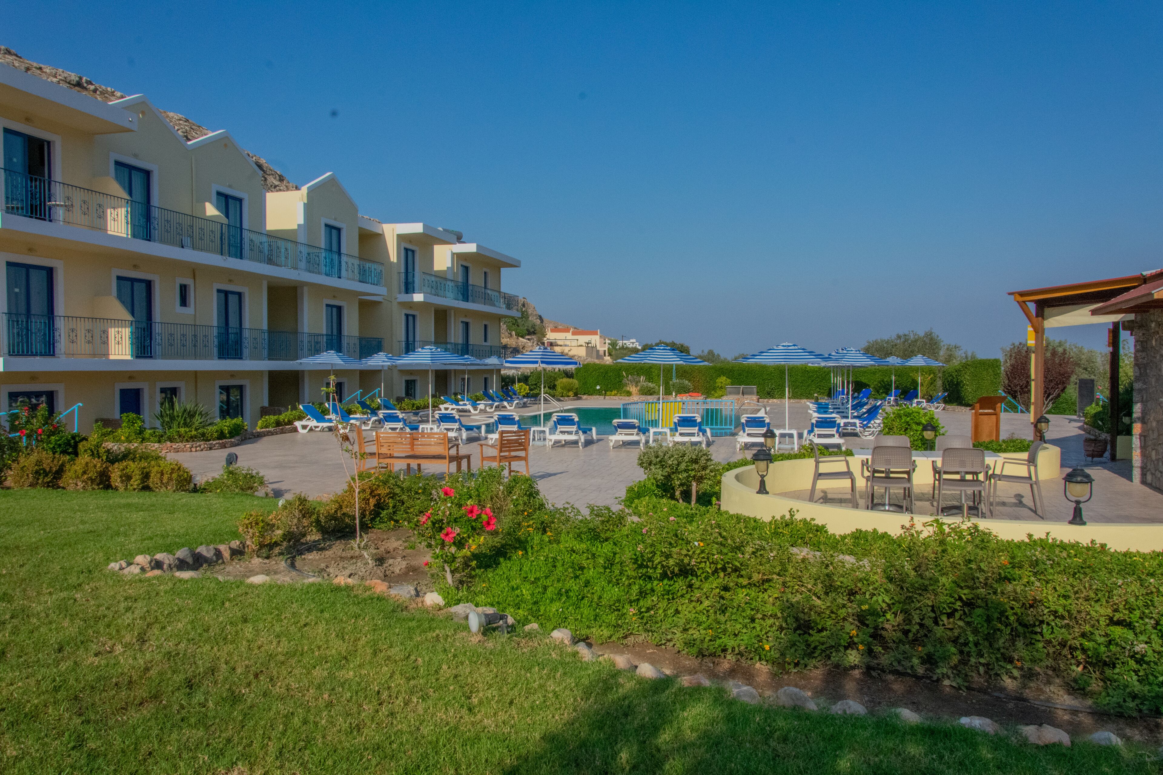 A hotel with multiple balconies, a pool area with sun loungers, and a well-maintained garden.