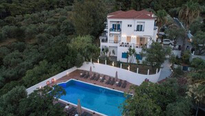 Una casa de dos pisos con piscina, rodeada de árboles y un muro blanco.