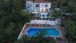 Un edificio de varios pisos con piscina, rodeado de árboles y una cerca blanca.