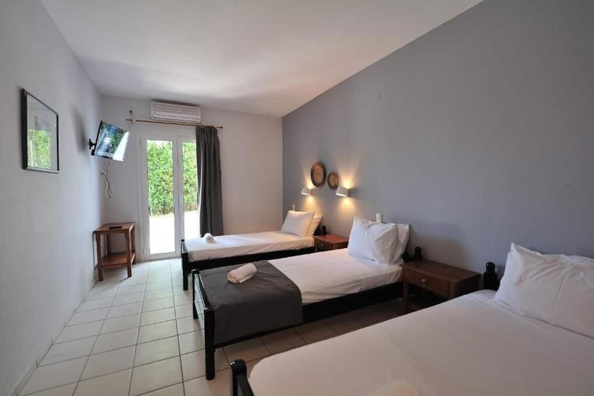 A hotel room with two single beds, a small wooden table, a TV mounted on the wall, and a sliding glass door leading to a balcony.