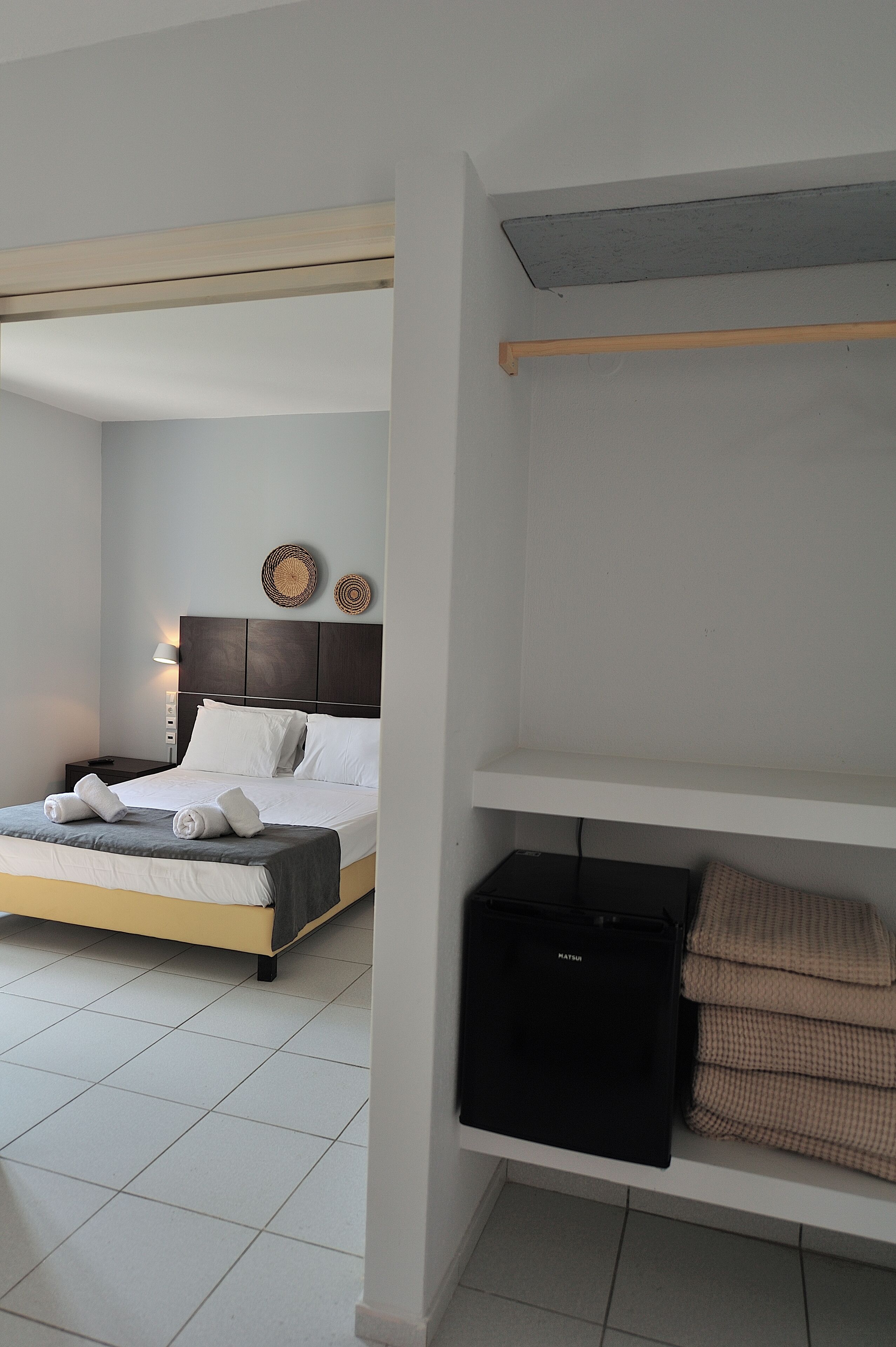 A hotel room with a bed, a nightstand, a lamp, a shelf with folded towels, and a black suitcase.
