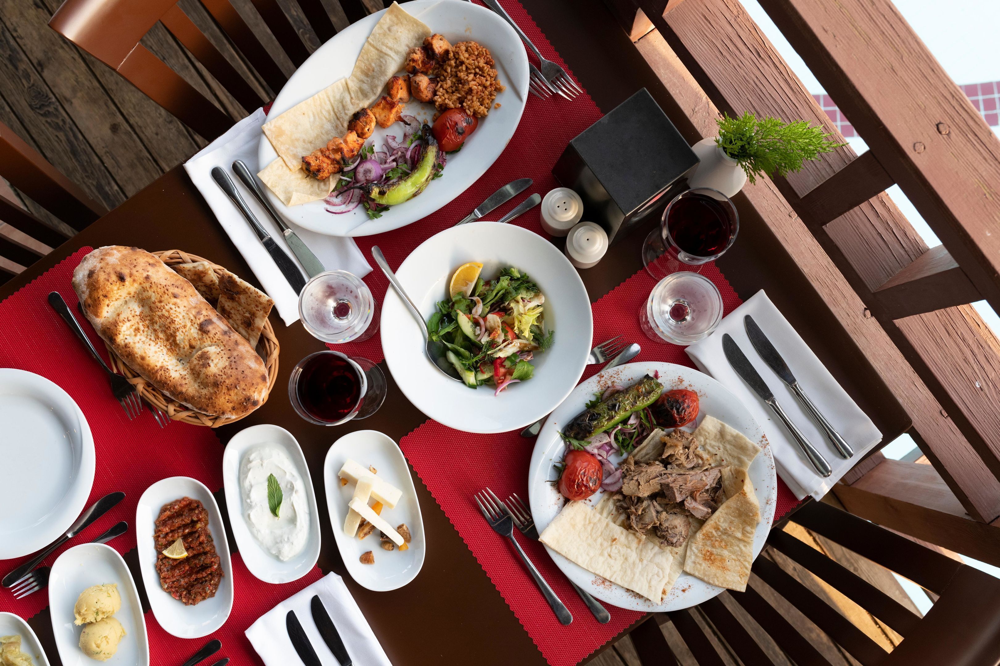 A table set with various dishes, including a plate of meat, a salad, and a bowl of yogurt, along with glasses of red wine.