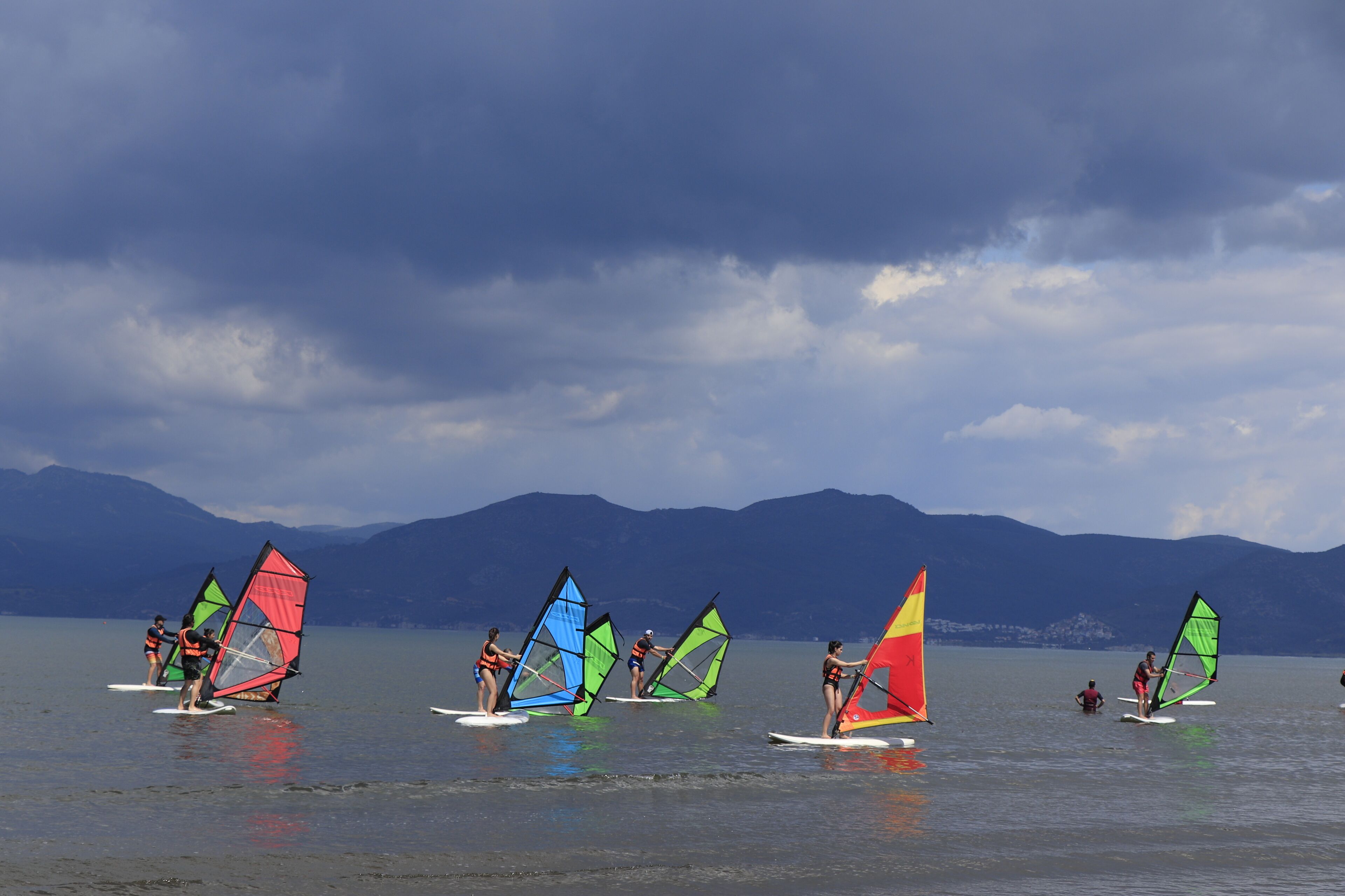 Eine Gruppe von Personen windsurft auf einem See vor einer Bergkulisse.