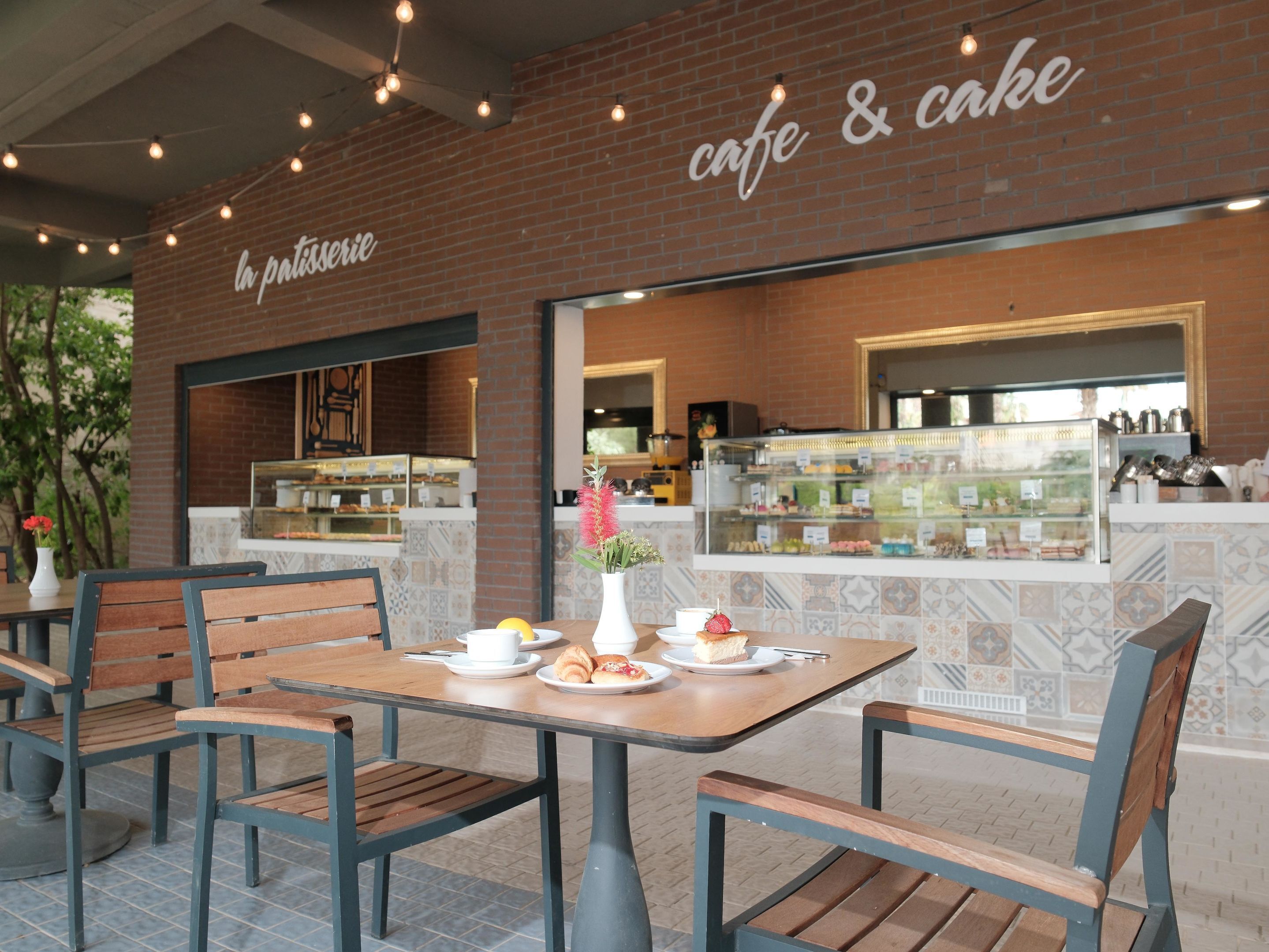 Ein Straßencafé mit Holztischen und -stühlen, einer Backsteinmauer und einer Vitrine mit Gebäck.