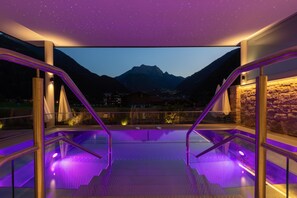 A modern outdoor pool area with glass railings and purple lighting, overlooking a mountainous landscape. - St. George (Mayrhofen)