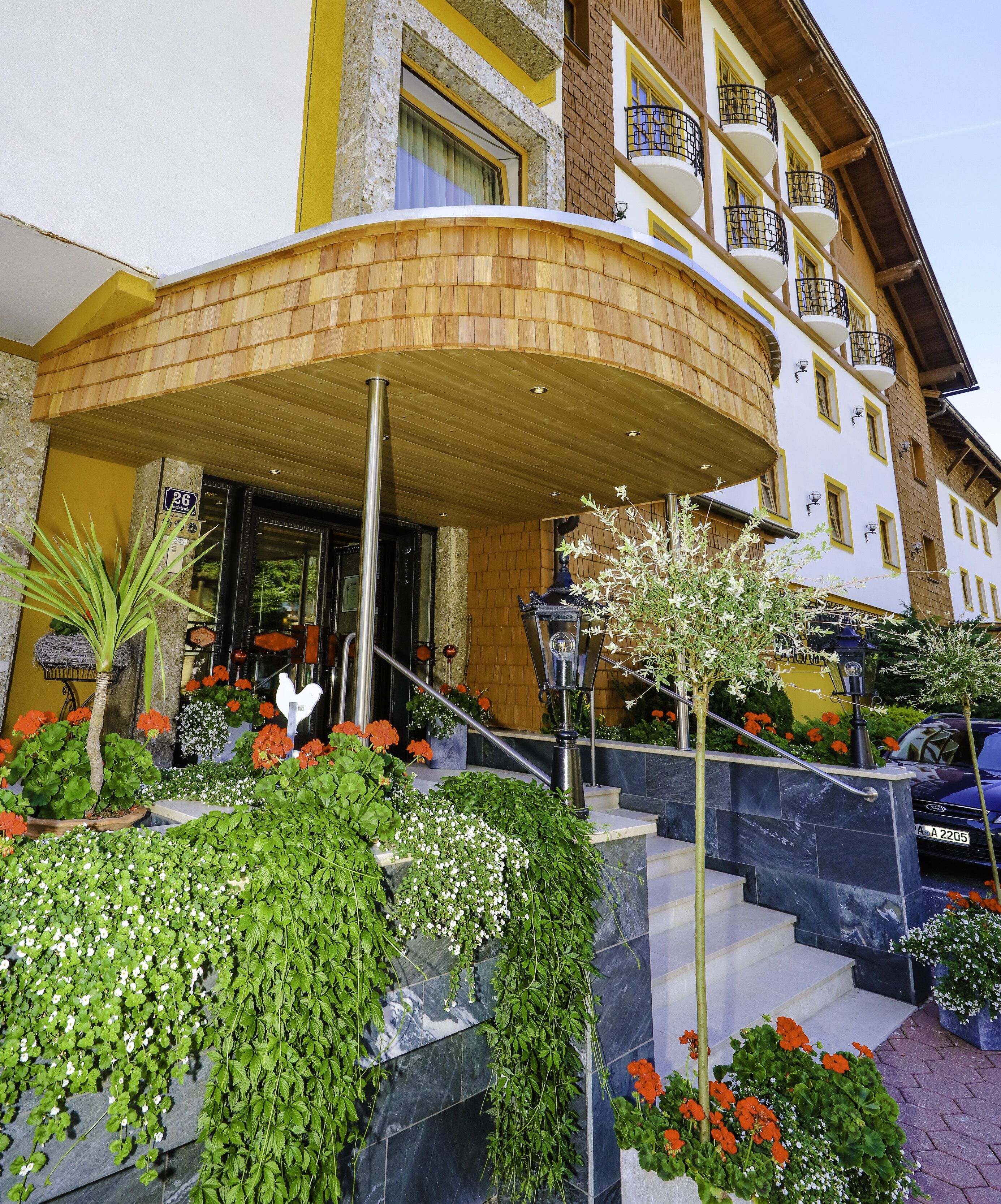 A hotel entrance with a curved wooden canopy, a staircase with marble steps, and a variety of potted plants and flowers.