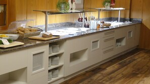 Montaje de bufé con diversos alimentos y condimentos sobre una encimera de mármol, rodeado de armarios blancos y un piso de madera.