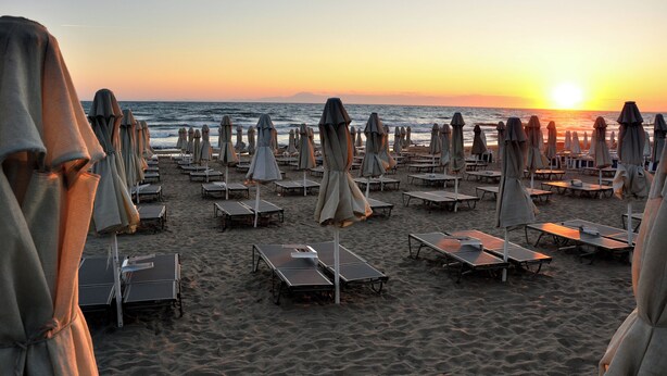 Uma praia com fileiras de espreguiçadeiras e guarda-sóis, alguns cobertos com panos, e um pôr do sol ao fundo.