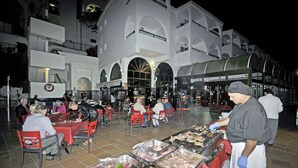 Un espace extérieur aménagé pour les repas, avec plusieurs tables et chaises, un barbecue où sont préparées différentes sortes de viandes, et un bâtiment aux fenêtres en arc de cercle en arrière-plan.