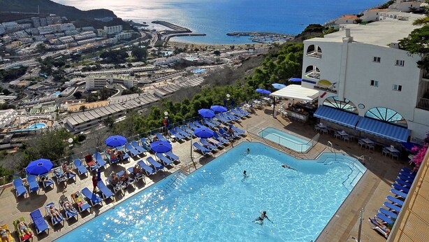 Une grande piscine, dotée de chaises longues et de parasols, entourée de bâtiments et offrant une vue sur la mer.