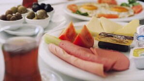 A table with plates of food including sliced tomatoes, cheese, olives, and a hot dog. There are also condiments and a glass of iced tea. - Palm Inn Hotel (Izmir)