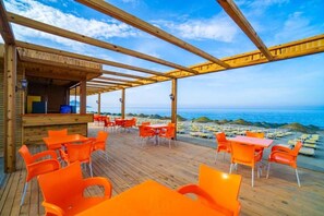 Une terrasse extérieure en bois, avec des chaises et des tables orange, donnant sur la mer.