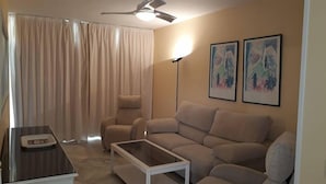A living room with a sofa, a coffee table, a chair, a ceiling fan, and two framed artworks on the wall. - Leo Las Americas (Isla Cristina)