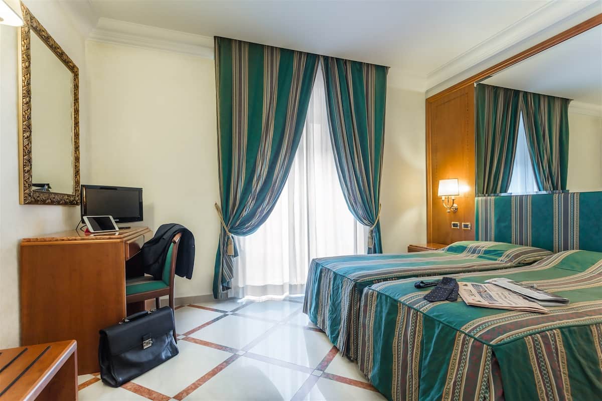 A hotel room with two beds, a desk with a computer, a chair, a large mirror, and a window with curtains.