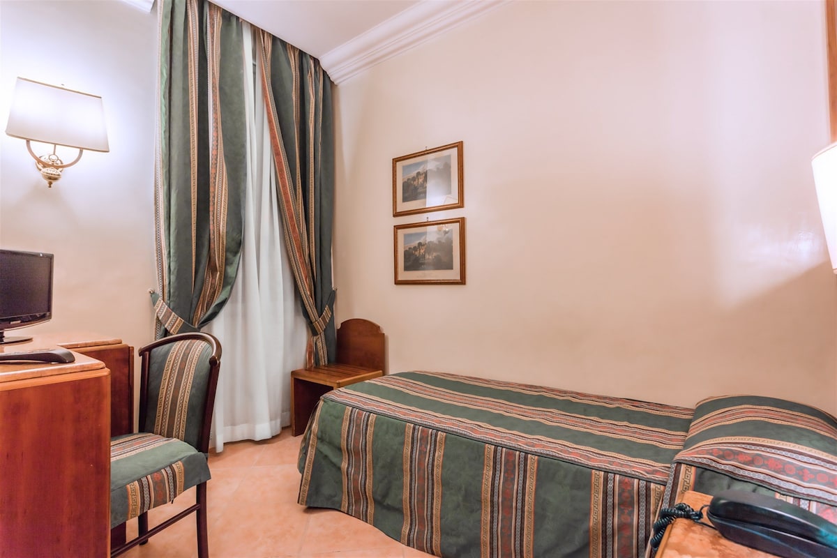 A hotel room with a bed, a desk with a computer, a chair, a lamp, and two framed pictures on the wall.