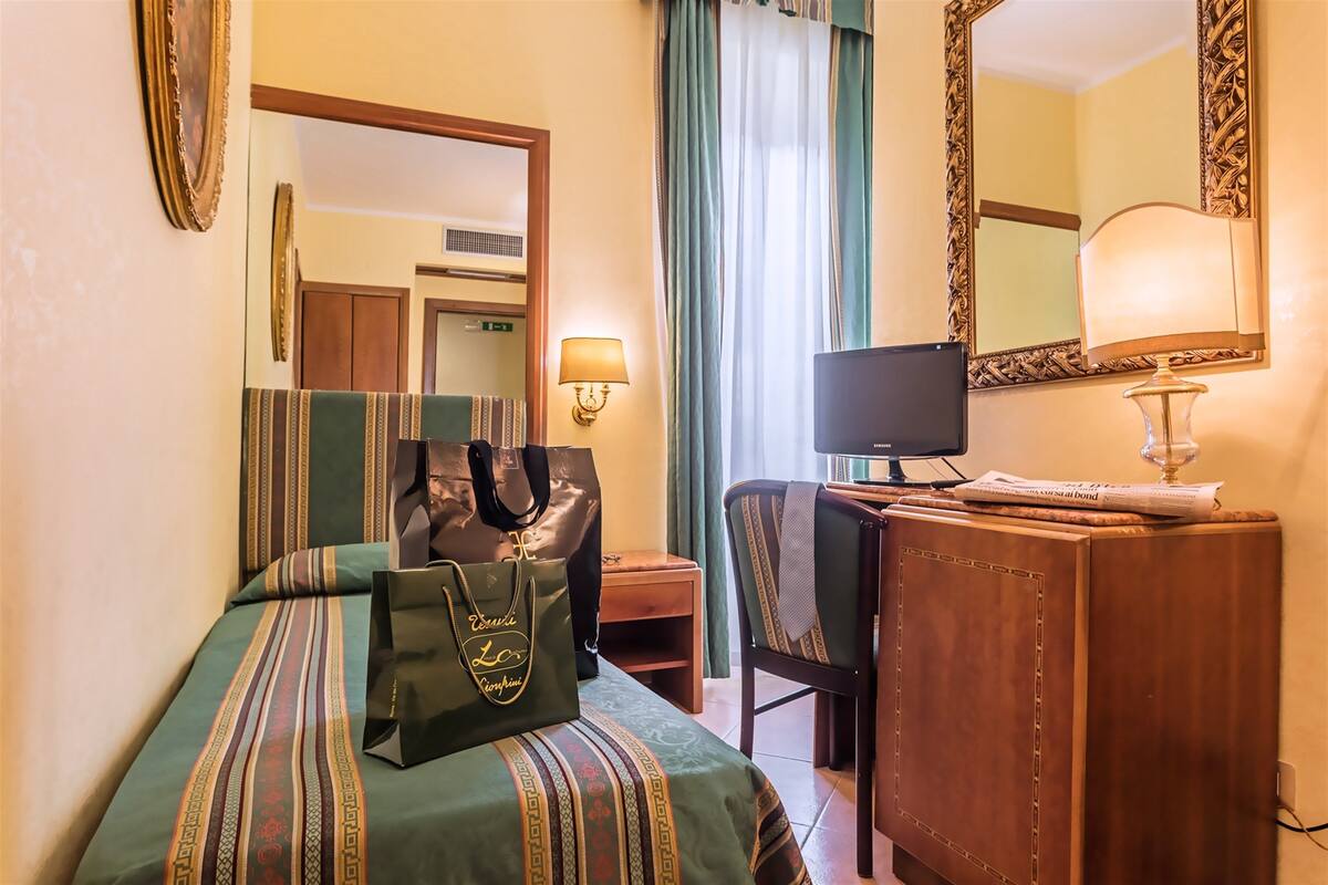 A hotel room with a bed, a desk with a computer, a chair, a lamp, and a large mirror.