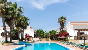 A swimming pool with lounge chairs, palm trees, and white buildings with red-tiled roofs. - Aparthotel Nuramar (Ciutadella de Menorca)