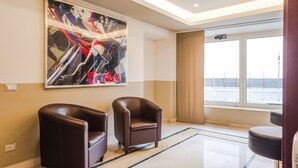 A hotel lobby with two leather armchairs, a large abstract painting, a large window with a view of a cityscape, and a modern ceiling design. - Raeli Hotel Siracusa (Rome)