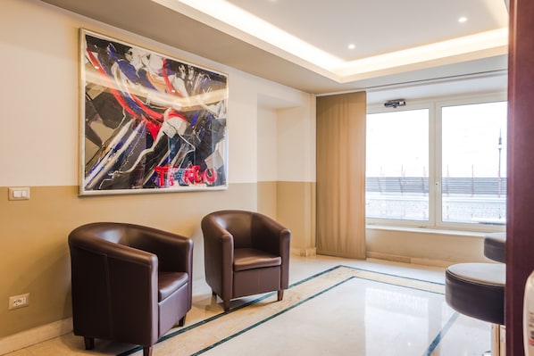 A hotel lobby with two leather armchairs, a large abstract painting, a large window with a view of a cityscape, and a modern ceiling design. - Raeli Hotel Siracusa (Rome)