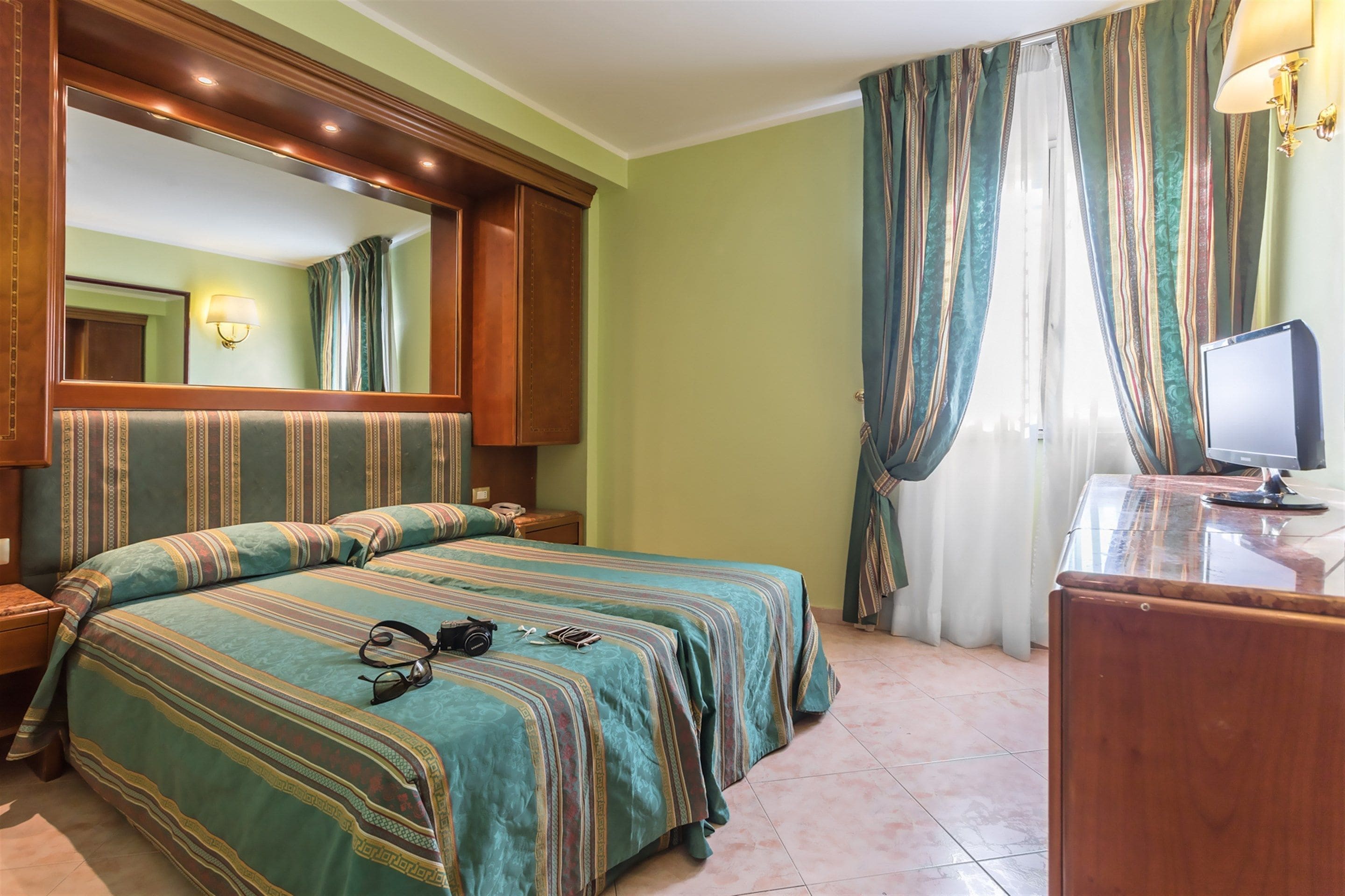 A hotel room with a large bed, a desk with a computer, a mirror, and curtains.