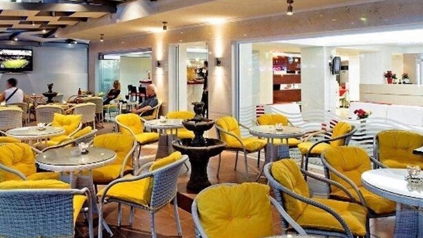 A restaurant with yellow chairs and tables, a fountain, and a TV on the wall. - Ambassador (Rhodes)