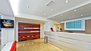 A modern reception area with a marble floor, a wooden bench, a reception desk, and a wall-mounted TV. - Ambassador (Rhodes)
