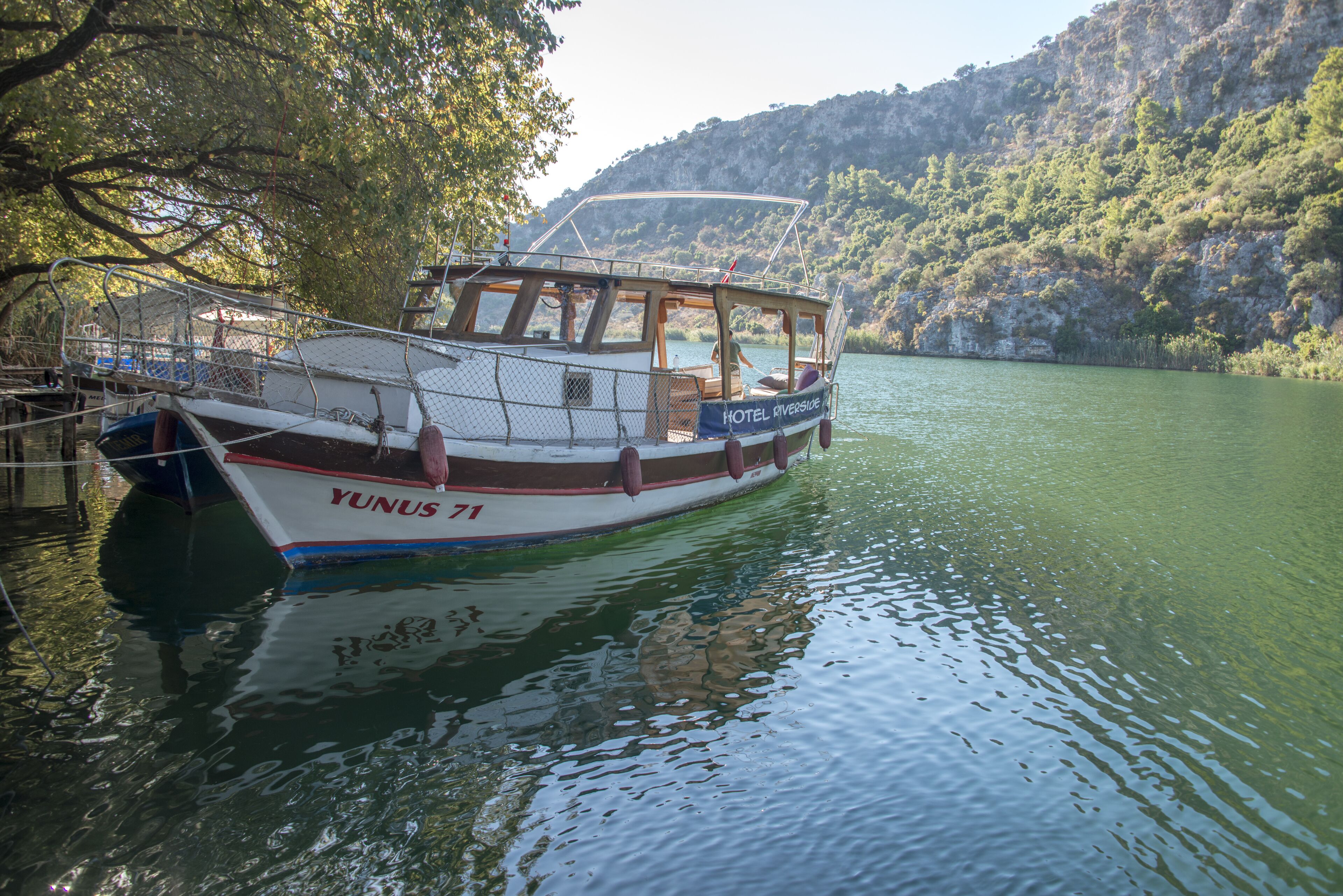 A boat named YUNUS 71 docked at a marina with a view of a hilly landscape.
