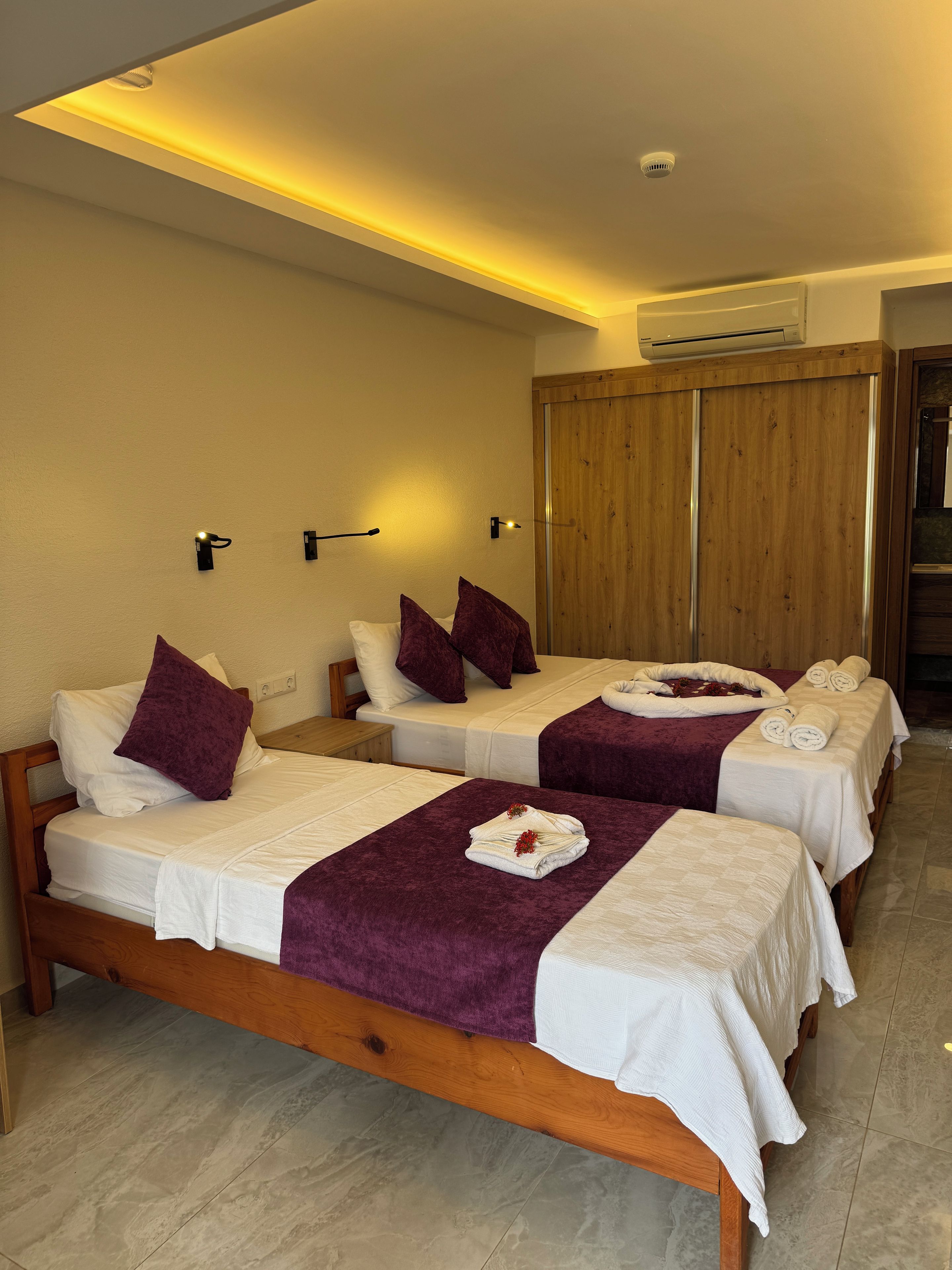 Una habitación de hotel con dos camas, cada una con una colcha morada y ropa de cama blanca, un armario de madera y un equipo de aire acondicionado en la pared.