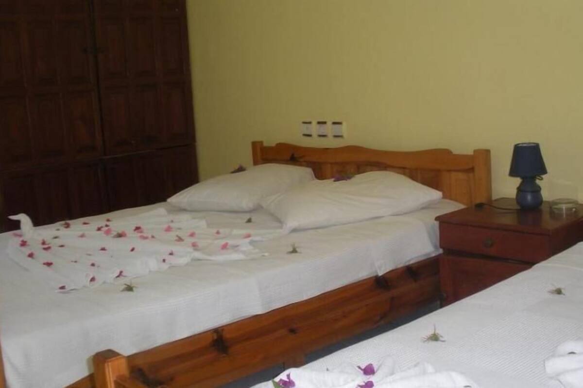 Una habitación con dos camas de madera, cada una con sábanas y almohadas blancas. Hay una mesita de noche de madera con una lámpara y un cajón pequeño.