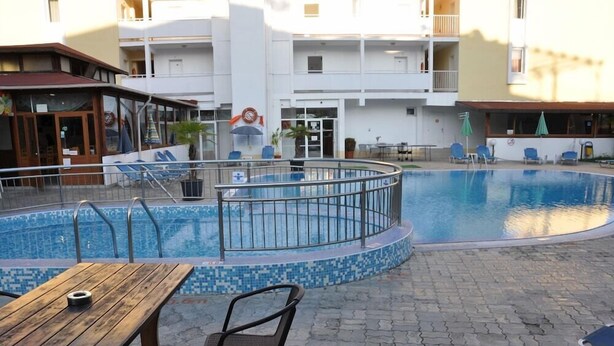 A pool area with a wooden table and chairs, a pool with a tiled edge, and a building with balconies in the background. - Hotel Belleville (Nessebar)