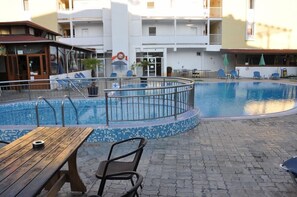 A pool area with a wooden table and chairs, a pool with a tiled edge, and a building with balconies in the background. - Hotel Belleville (Nessebar)