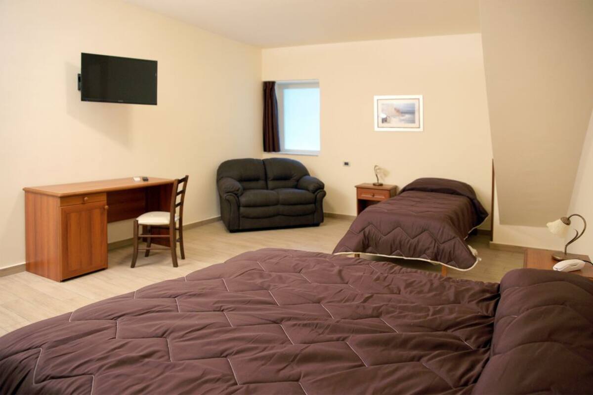 A bedroom with two beds, a desk, a chair, a TV, a window, and a bedside table with a lamp.