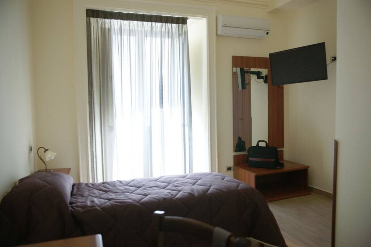 A hotel room with a bed, a chair, a desk, a television mounted on the wall, and a window with curtains.