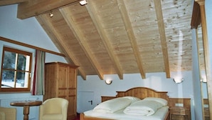 Un dormitorio de madera con techo inclinado, una cama, una silla, una mesita y una ventana.