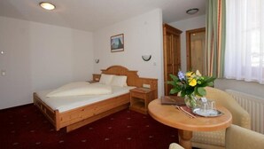 Habitación de hotel con una cama de madera, una mesita de noche pequeña, una silla, una mesa con un florero y una ventana con cortinas.