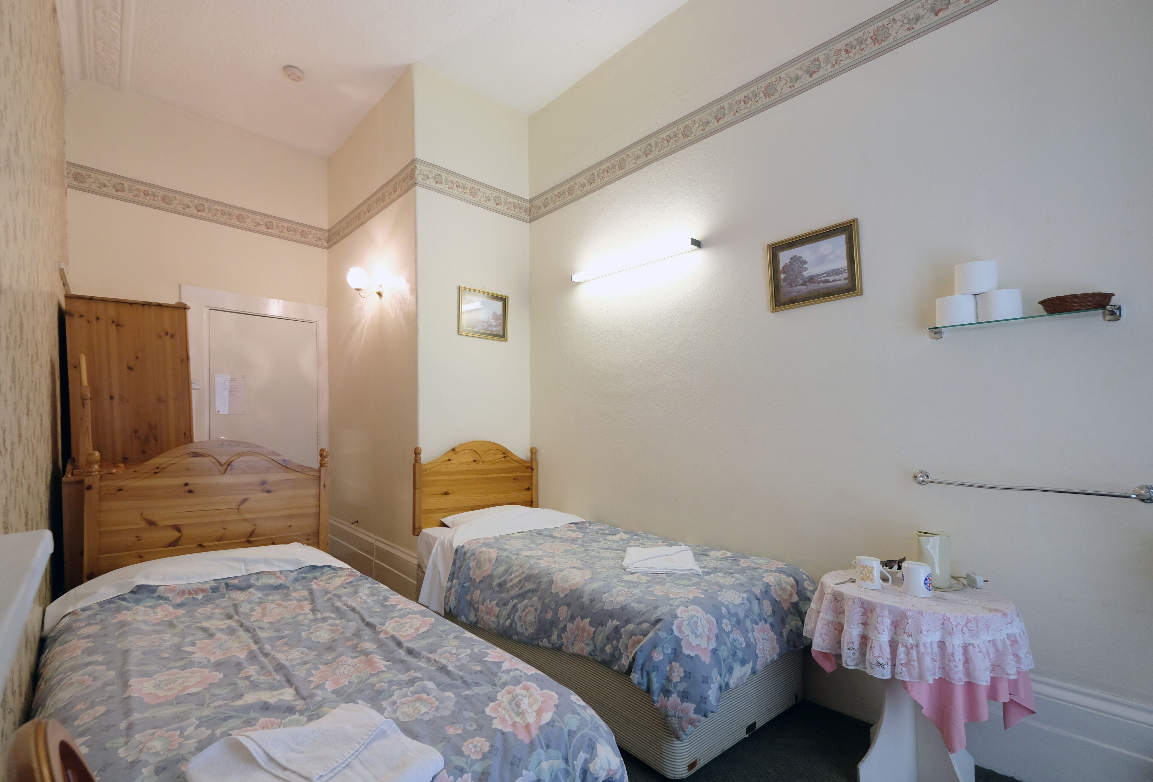 Une chambre d’hôtel avec deux lits en bois, une petite table recouverte d’une nappe rose et une étagère avec des articles de toilette.