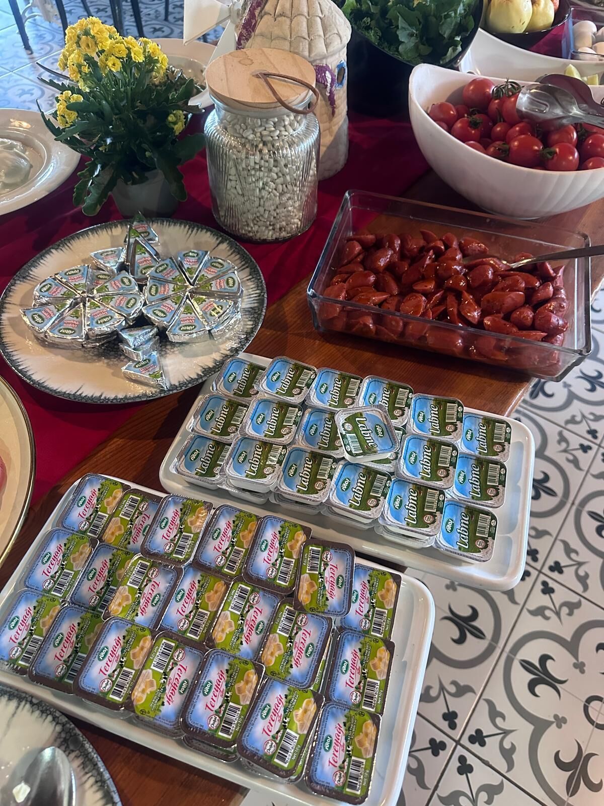 A table with various food items including a tray of packaged yogurt, a bowl of cherry tomatoes, and a dish of sausages.