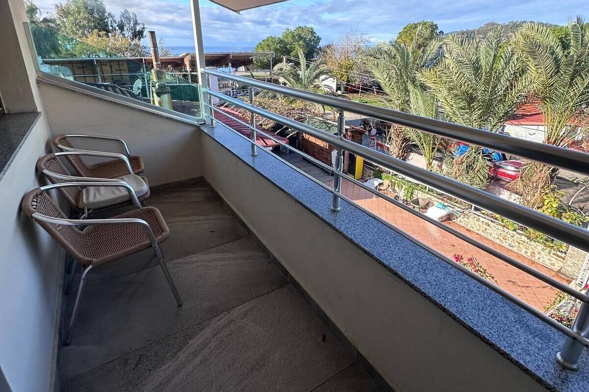 A balcony with a metal railing, a few wicker chairs, and a view of a street with trees and buildings.