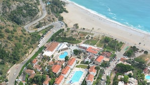 An aerial view of a coastal resort with multiple swimming pools, a beach, and a winding road. - The Kumsal Hotel Ölüdeniz (Fethiye)