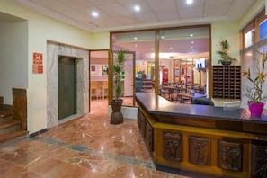 Un hall d’hôtel avec un comptoir d’accueil, une grande plante en pot et un escalier menant à un ascenseur.