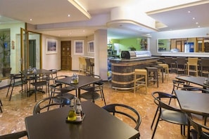 Un restaurante de hotel con suelo de mármol, varias mesas de comedor, sillas, una barra con máquina de café y cuadros en las paredes.
