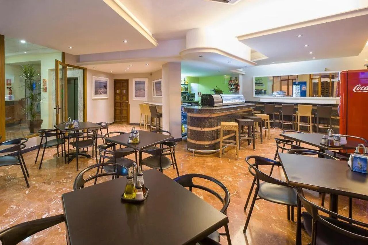 A hotel restaurant with multiple dining tables and chairs, a bar area, and a Coca-Cola refrigerator.