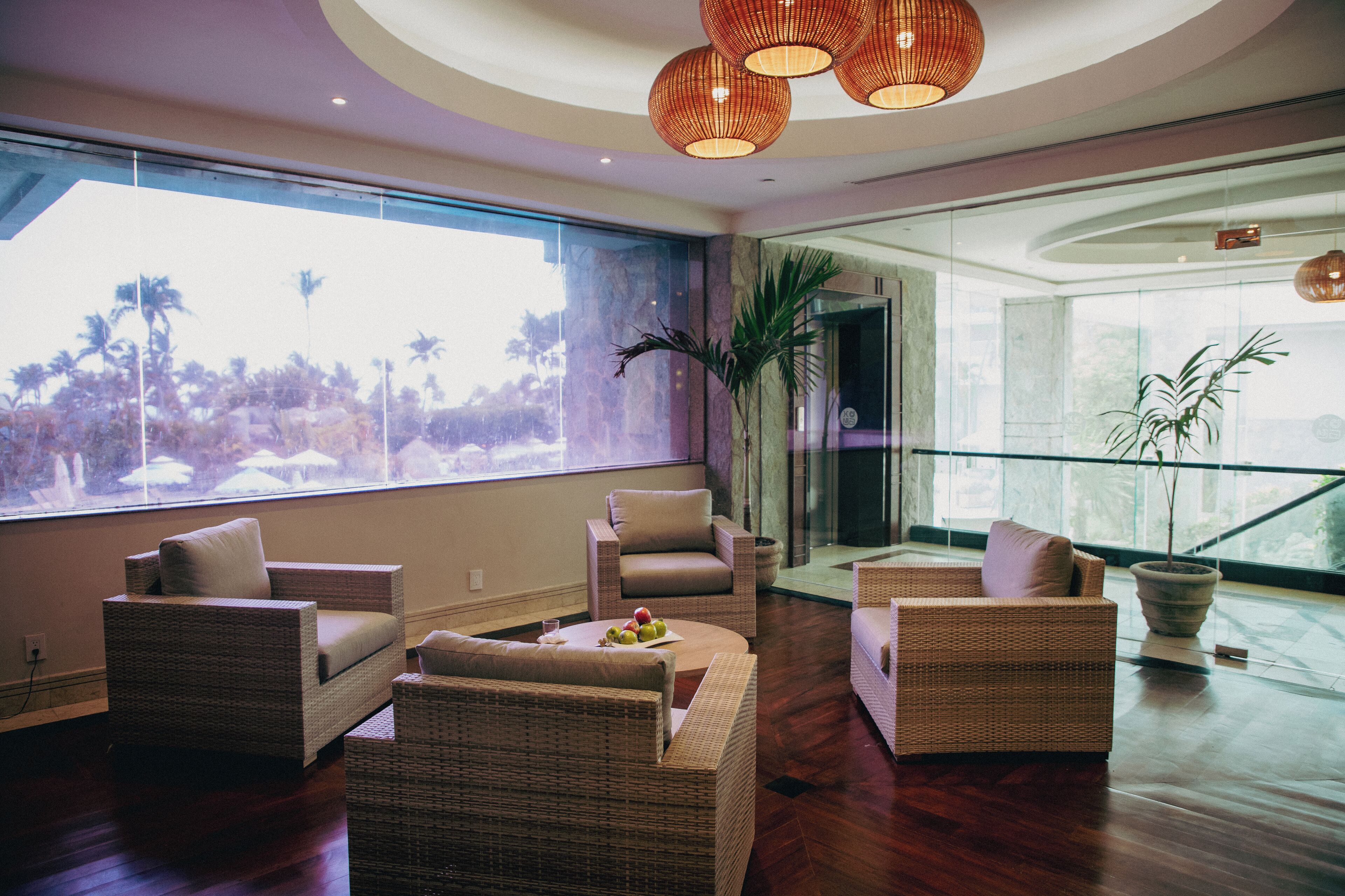 A spacious living area with wicker furniture, large windows, and a view of palm trees.