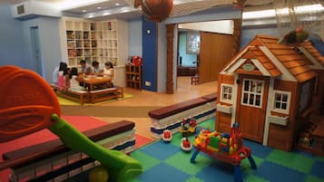 Zona de juegos infantiles con una casita, una mesa donde niños juegan y estantes con juguetes.