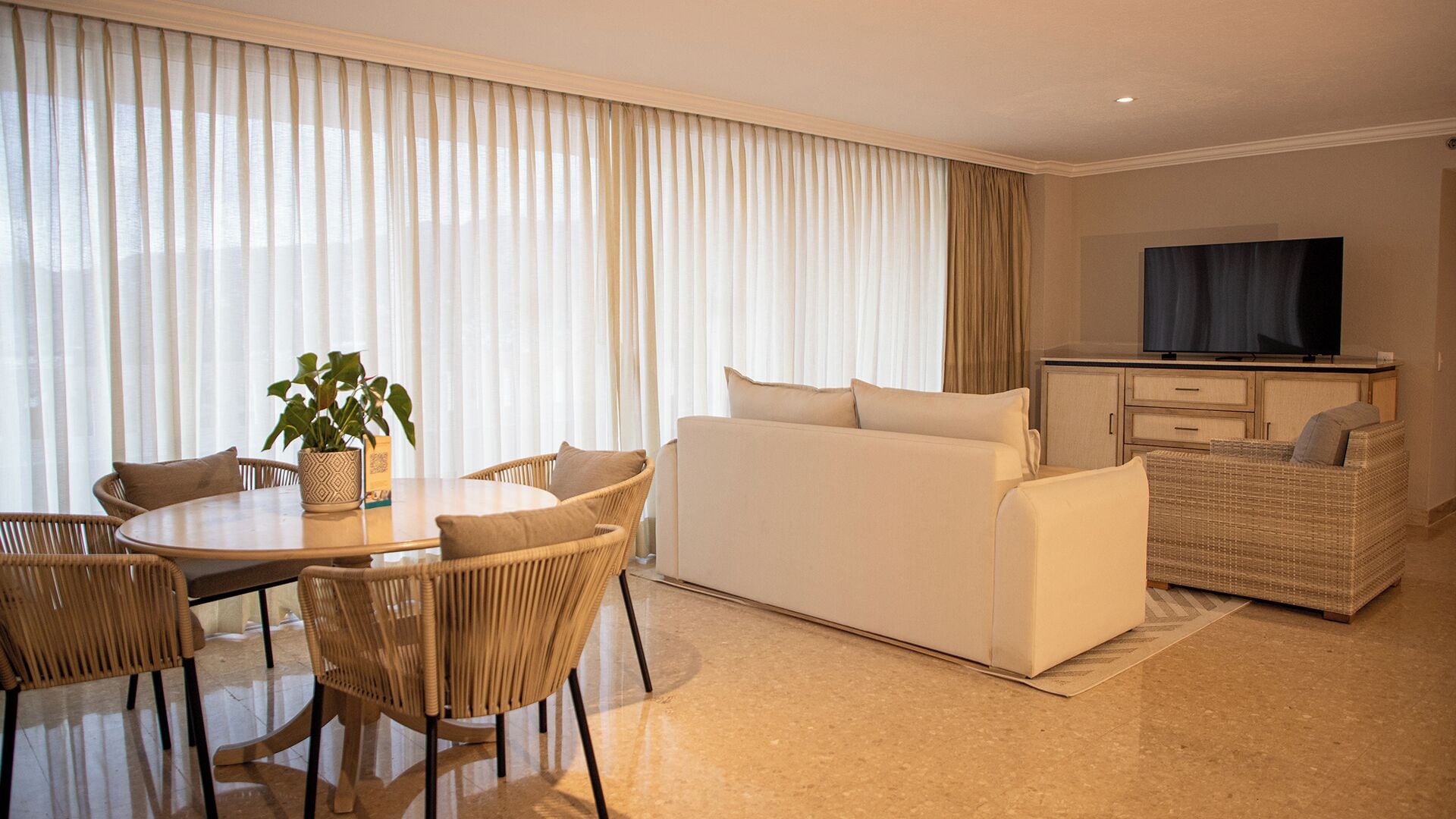 A modern living room with a dining area, a sofa set, a television, and a wicker chair.