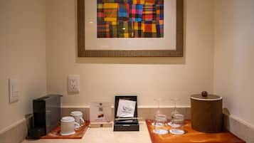Una habitación pequeña con un cuadro abstracto enmarco, una estación de café con máquina Nespresso, tazas y vasos, y una cesta decorativa.