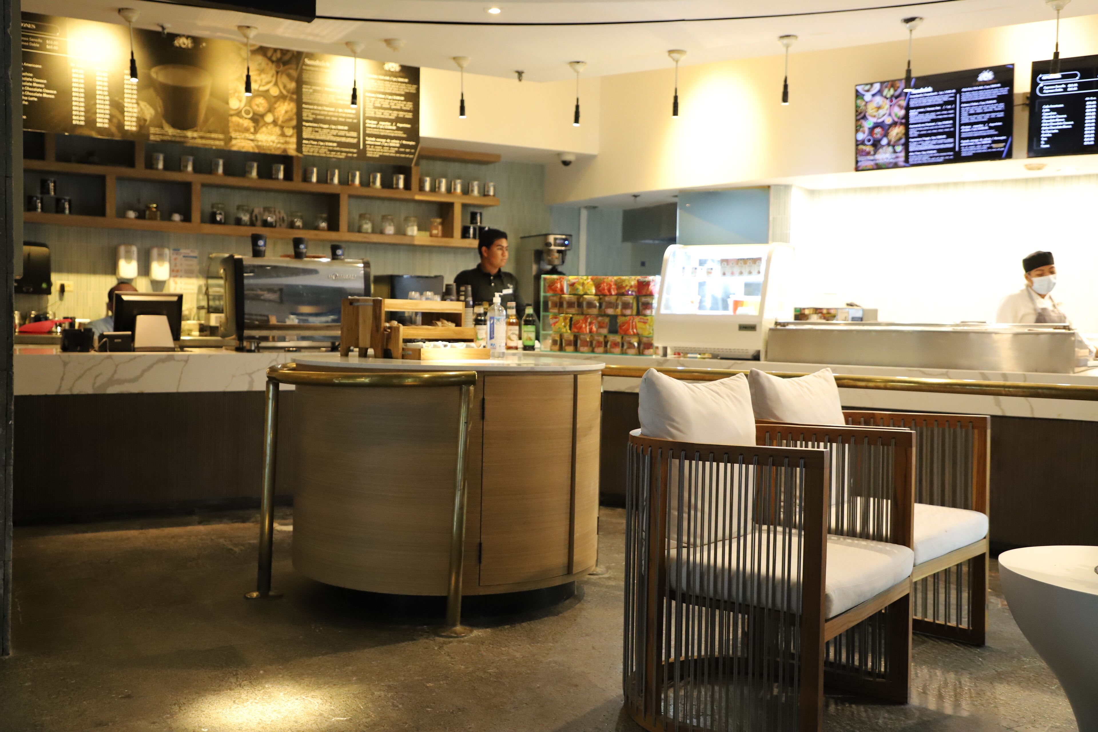 Interior moderno de un café con barra, mesas y una pantalla digital con el menú.