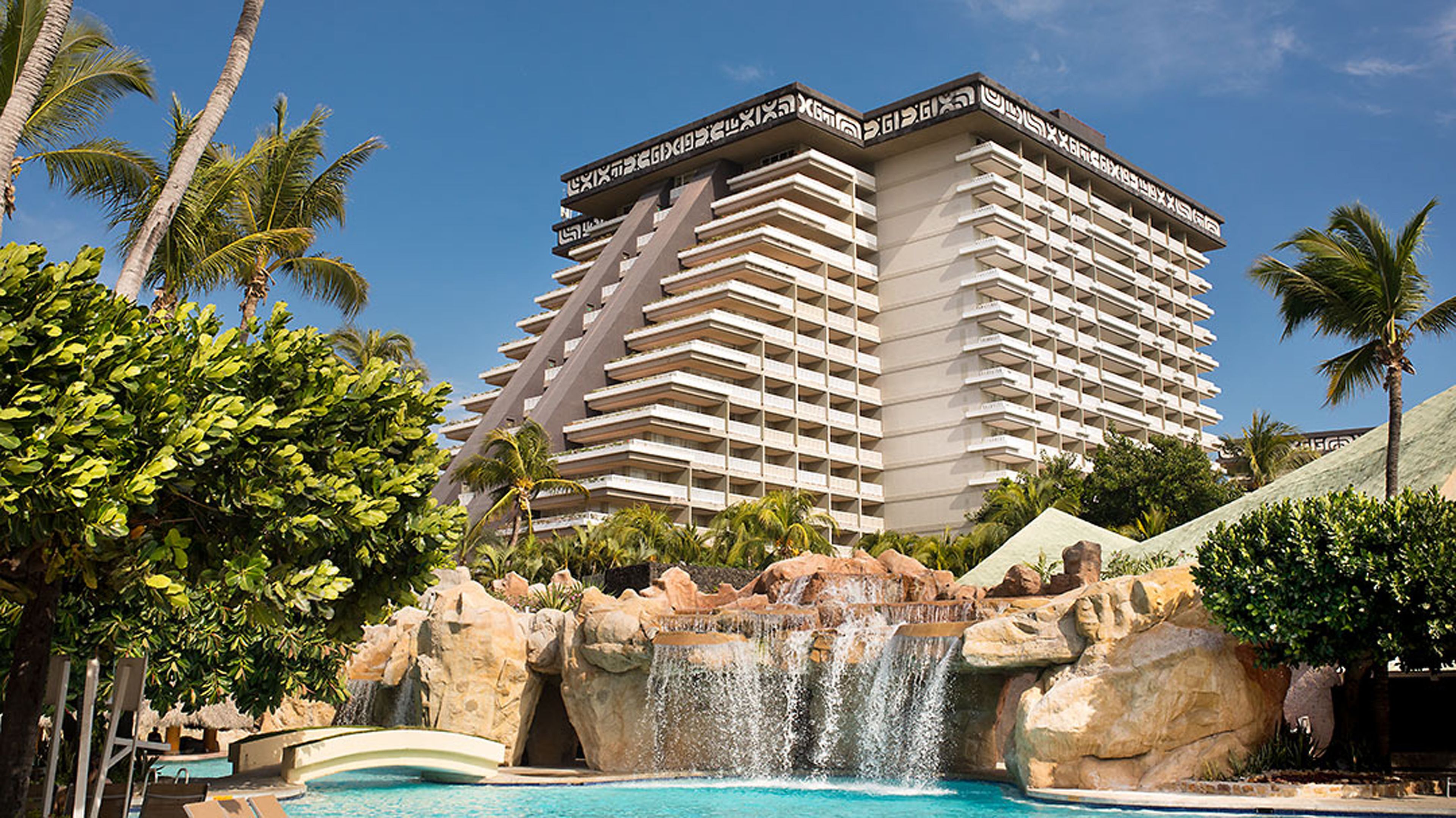 A hotel with a modern design, featuring a large pool with a waterfall, surrounded by palm trees and a rocky landscape.
