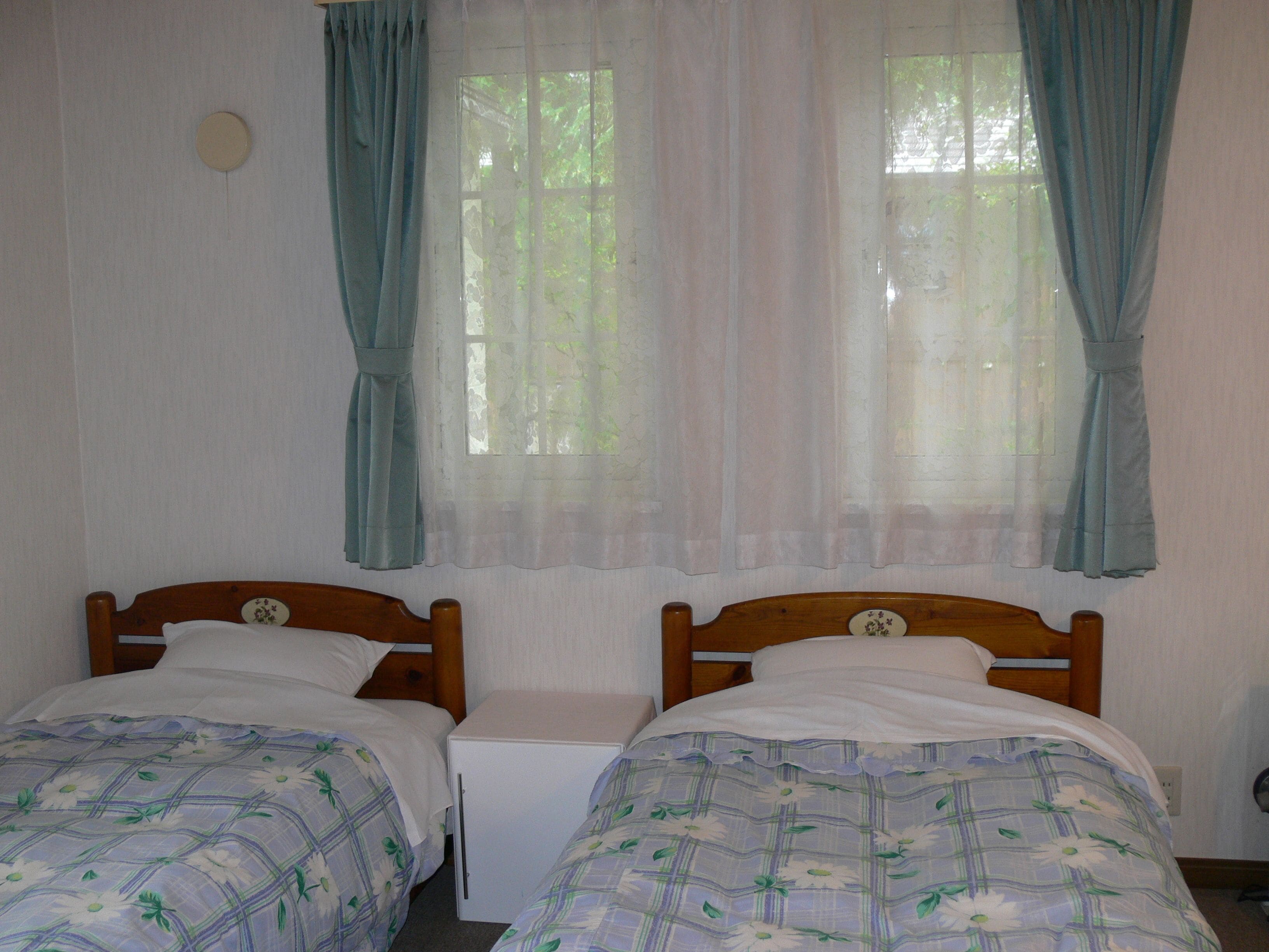Un dormitorio con dos camas individuales, una mesita de noche pequeña y una ventana con cortinas traslúcidas.
