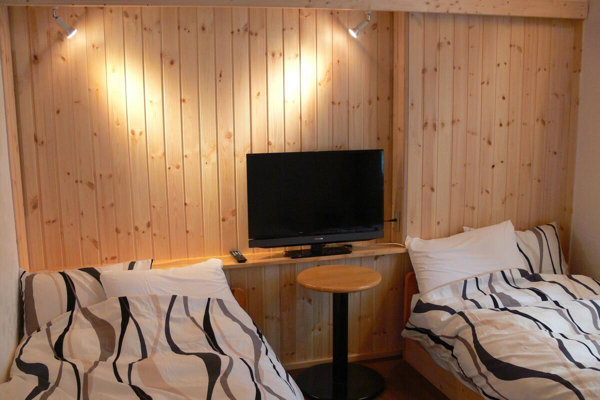 Ein Zimmer mit Holzwandverkleidung, einem Fernseher auf einem Standfuß, zwei Betten mit gestreifter Bettwäsche und einem kleinen runden Tisch.